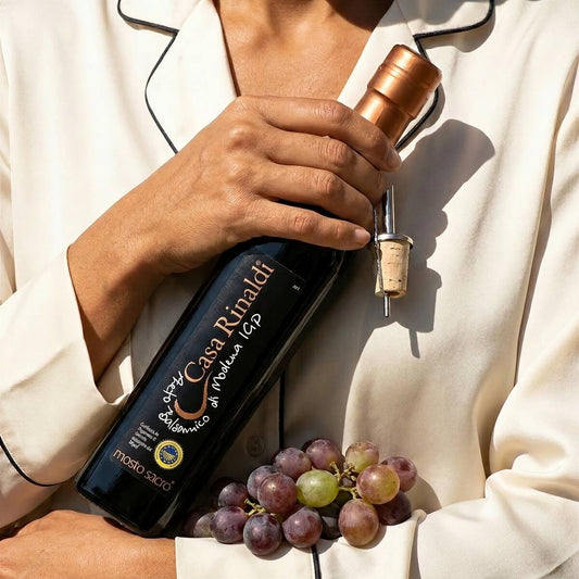 Person holding a bottle of Casa Rinaldi Mosto Sacro Balsamic Vinegar of Modena PGI, 250ml, with fresh grapes.