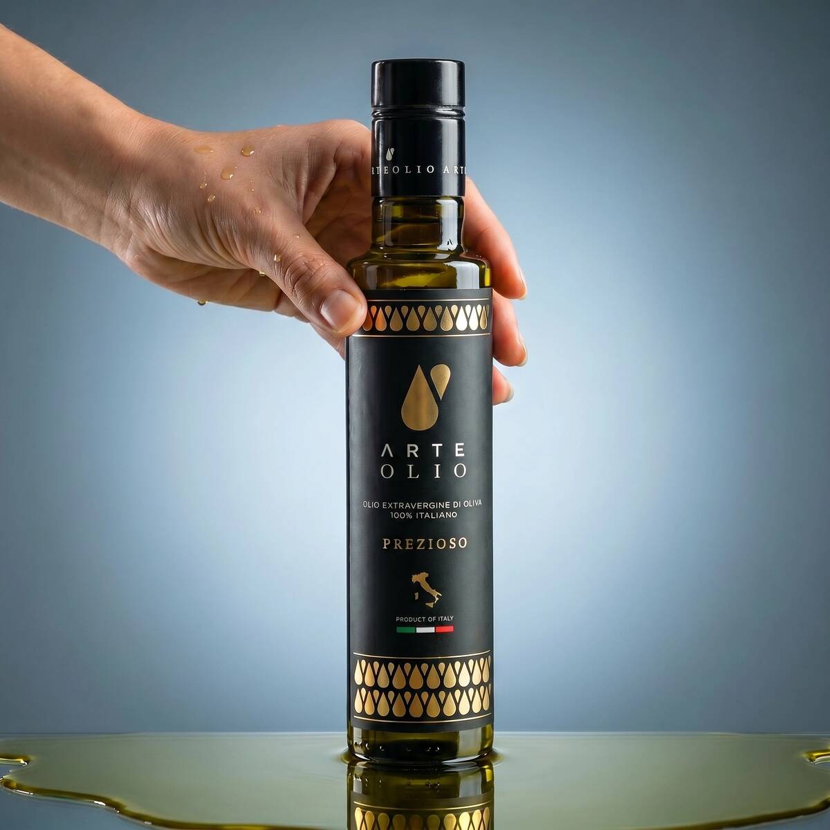 Hand holding Arte Olio Prezioso 100% Italian Extra Virgin Olive Oil 500ml bottle with olive oil pooling beneath on a grey background