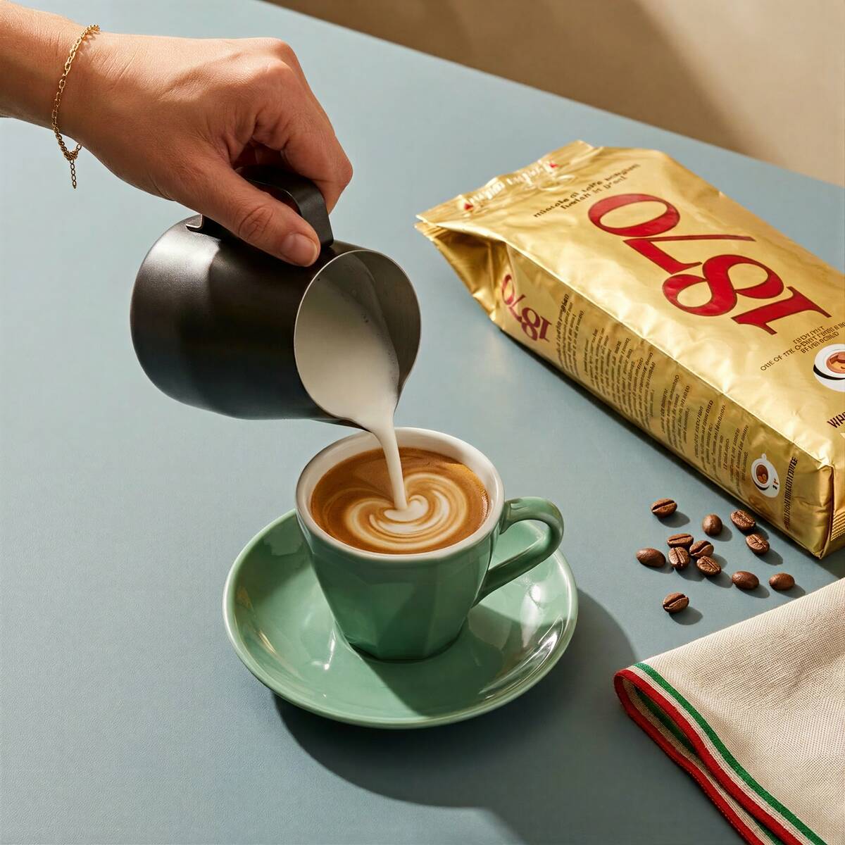 Hand pouring steamed milk into a latte art cappuccino next to a Caffè Barbera 1870 Mago whole bean coffee bag and scattered coffee beans