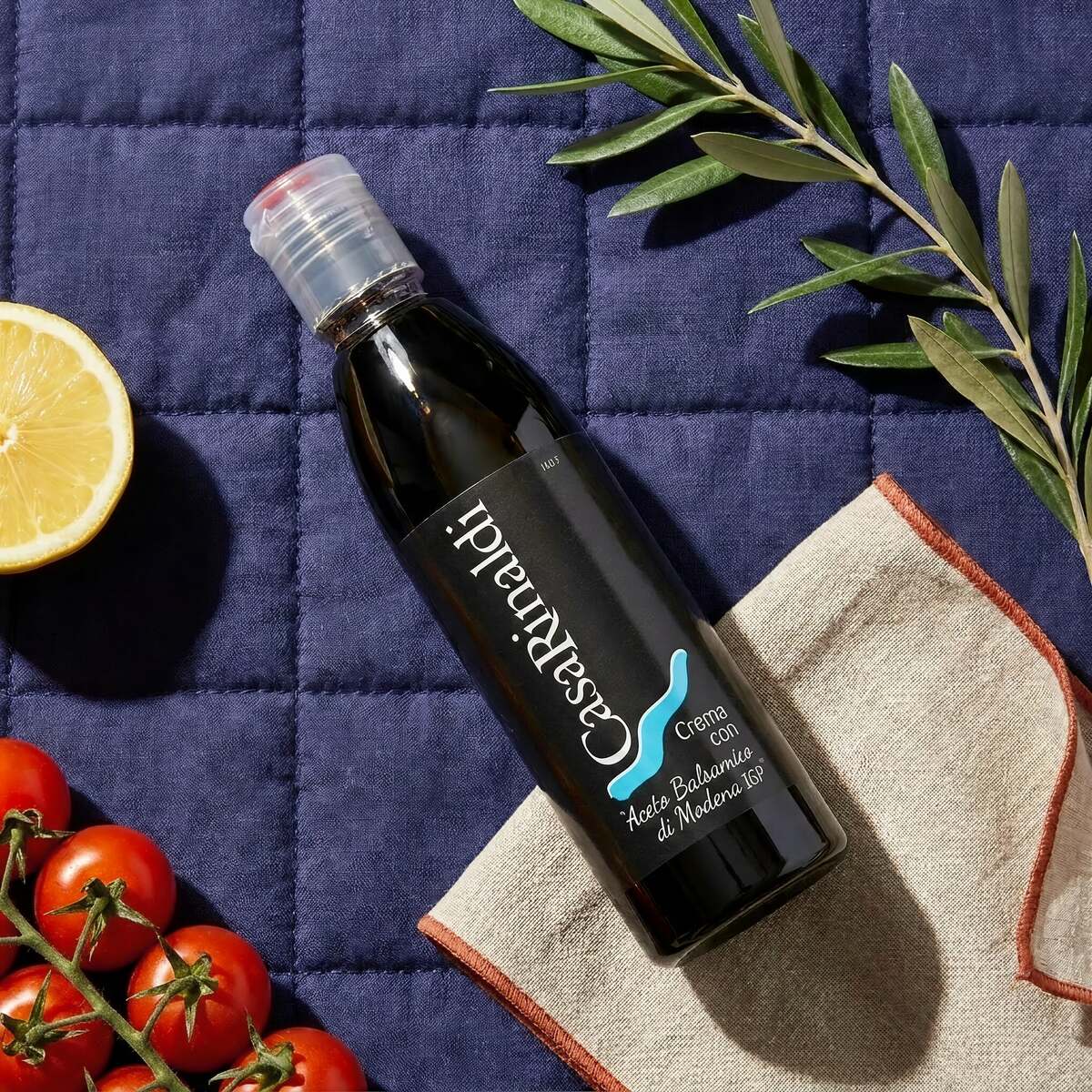 Casa Rinaldi Balsamic Glaze of Modena PGI 250ml bottle on a navy quilted cloth with cherry tomatoes, lemon, and olive branch