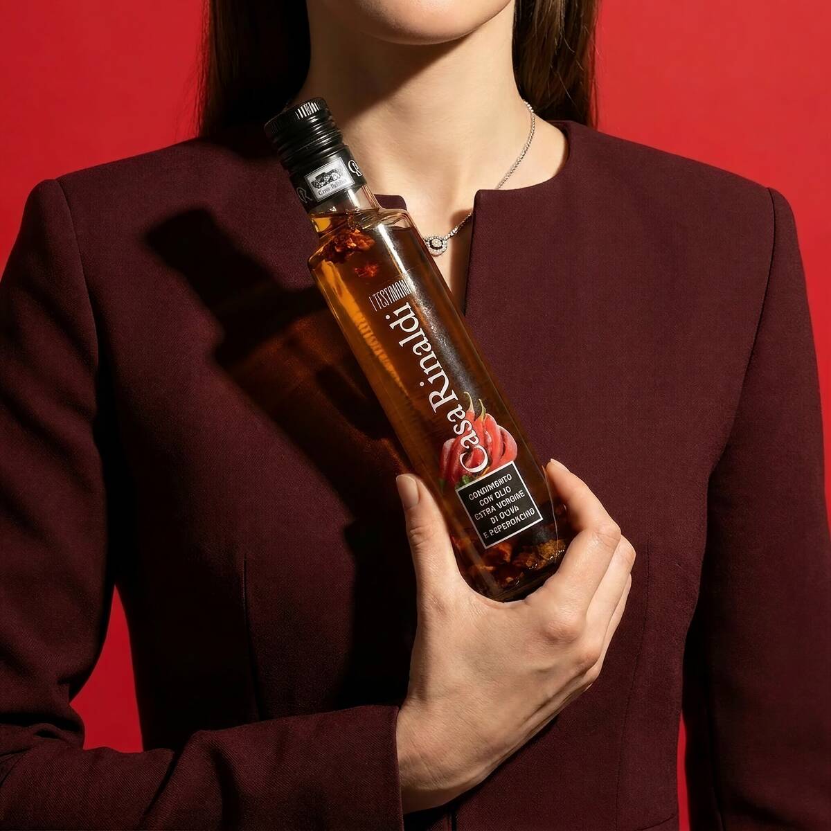 Woman in burgundy blazer holding a bottle of Casa Rinaldi I Testimoni Chili Flavored Extra Virgin Olive Oil 250ml against a red background
