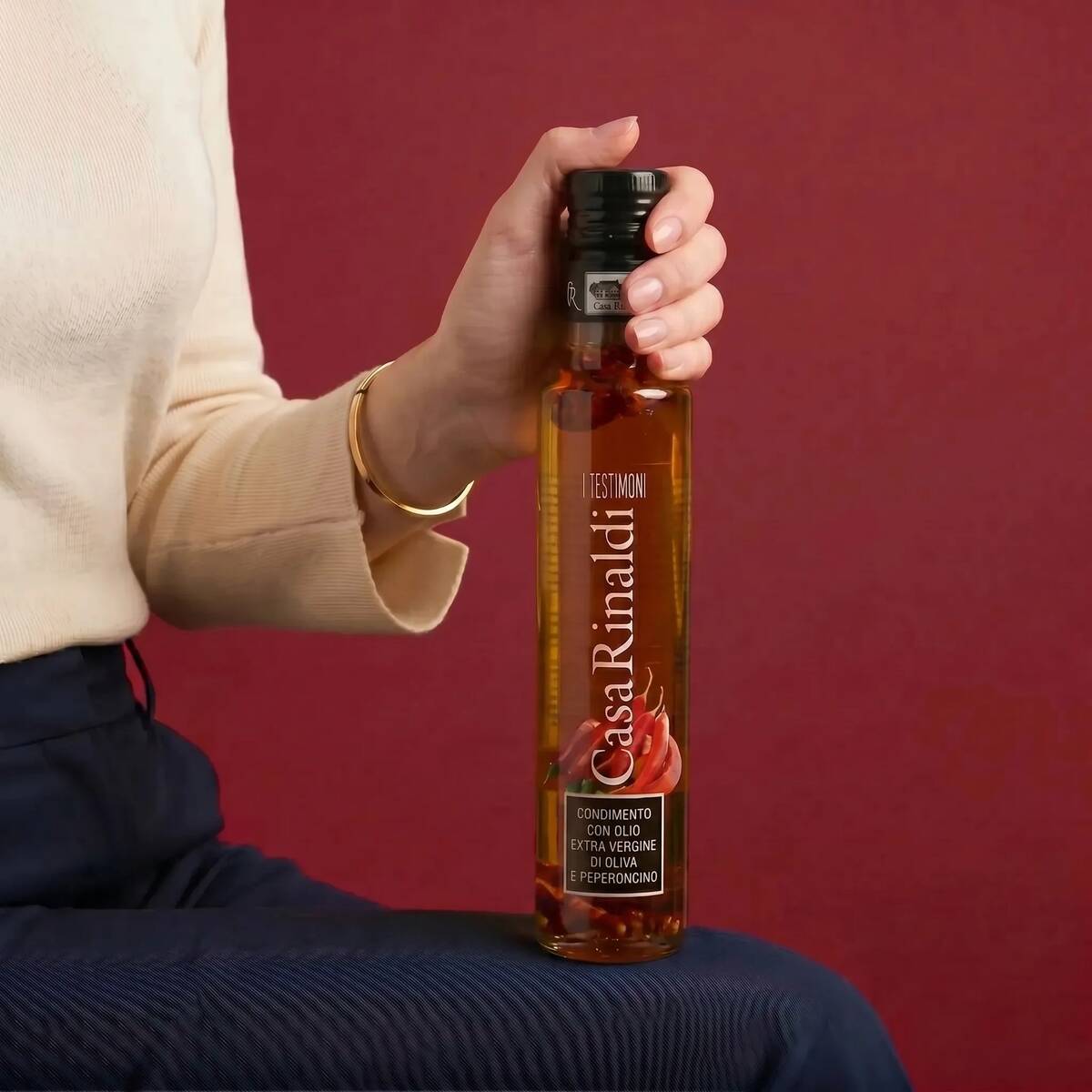 Hand holding a bottle of Casa Rinaldi I Testimoni Chili Flavored Extra Virgin Olive Oil 250ml against a deep red background