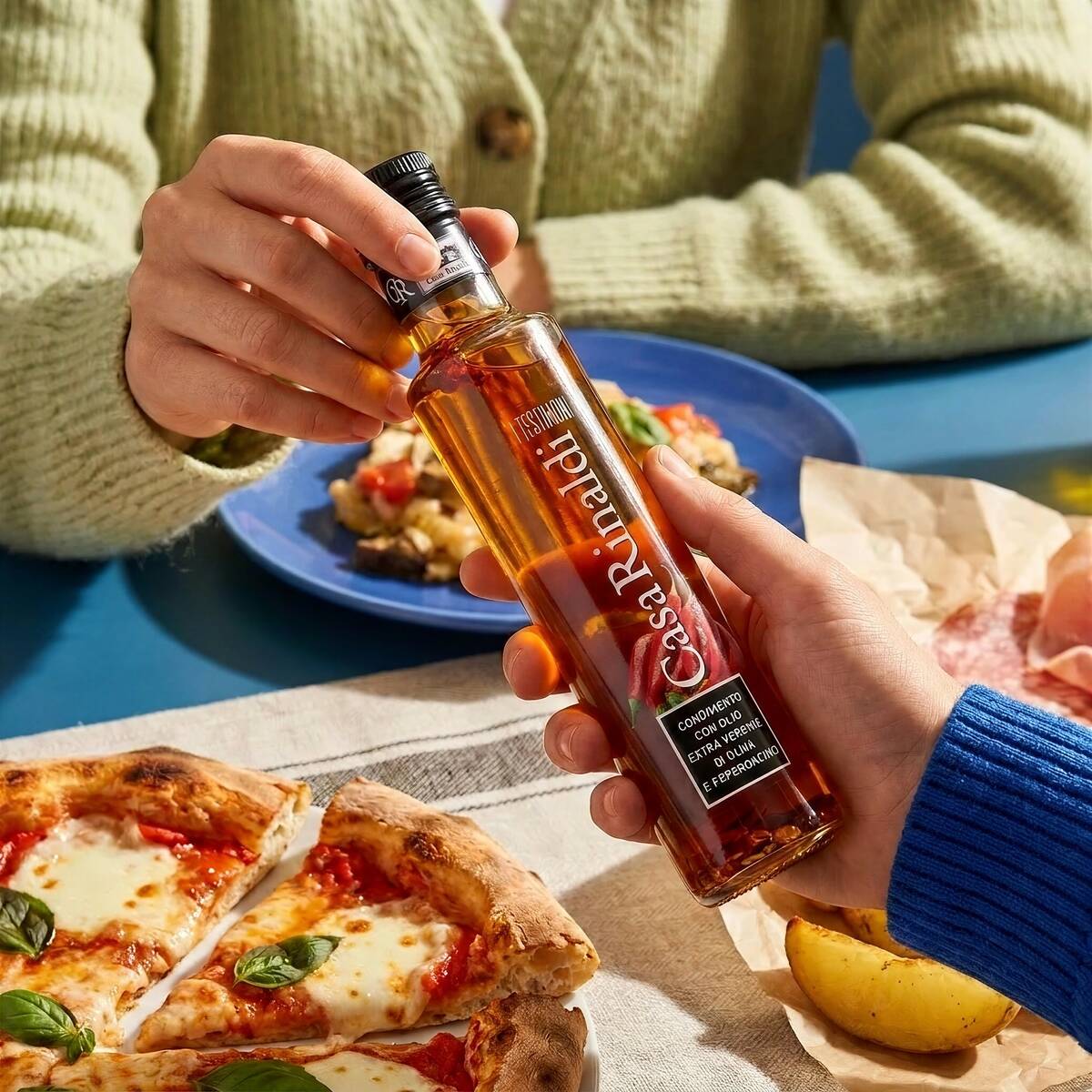 Two people passing a bottle of Casa Rinaldi I Testimoni Chili Flavored Extra Virgin Olive Oil 250ml over a Neapolitan pizza and charcuterie spread