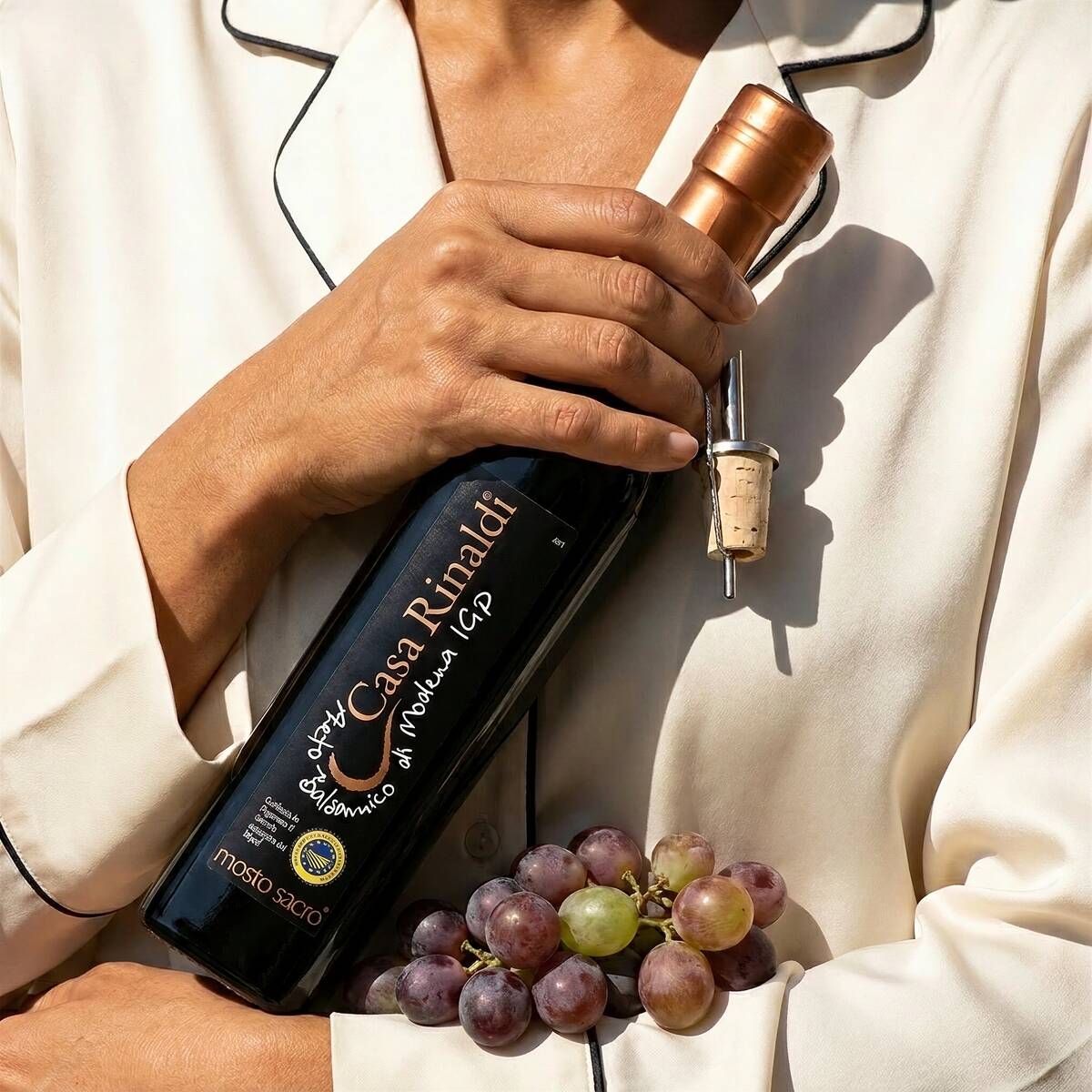 Person holding a bottle of Casa Rinaldi Mosto Sacro Balsamic Vinegar of Modena PGI, 250ml, with fresh grapes.